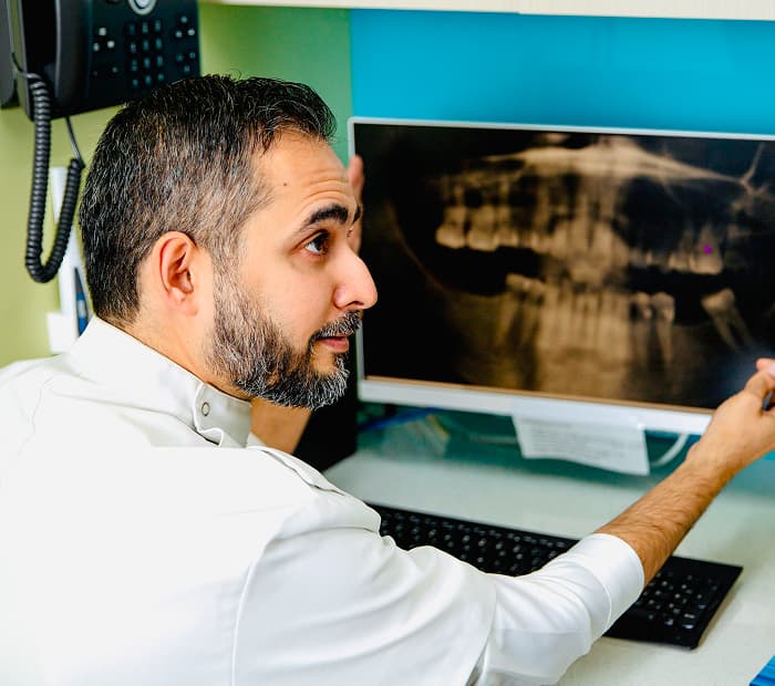 A person in a white clinical coat beside a monitor displaying a panoramic dental X-ray.