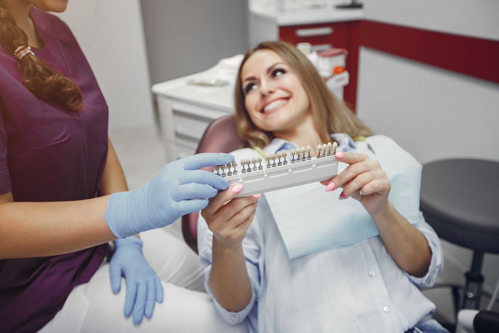Smiling female patient in a dental clinic holding a shade guide for tooth colour matching with a dental professional wearing gloves.